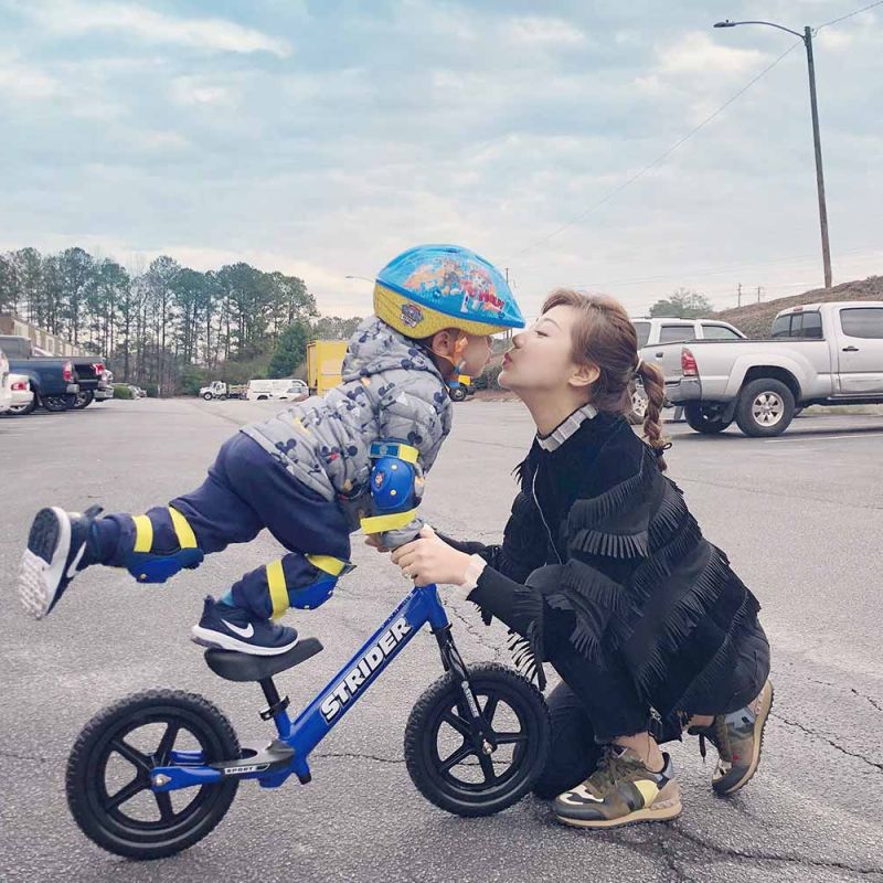 A child stands on the seat of a blue Strider 12 Sport to kiss mom