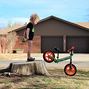 A boy stands on a stump while watching his Strider Bike roll away