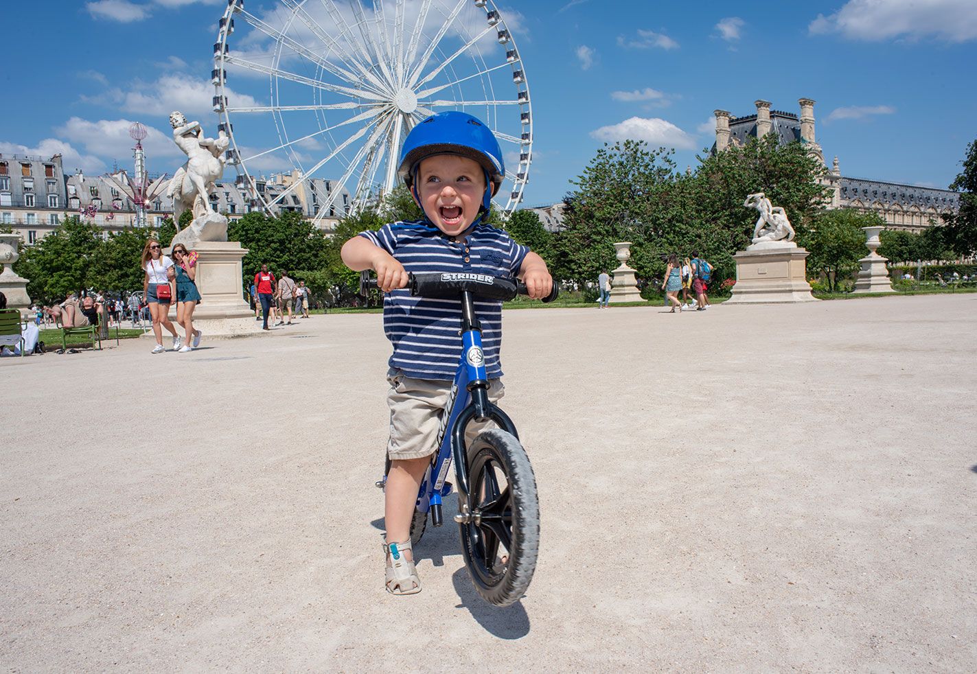 A little boy does a wheelie on his blue Strider 12 inch Sport balance bike in front of the ferris wheel outside the Tuileries Garden in Paris, France