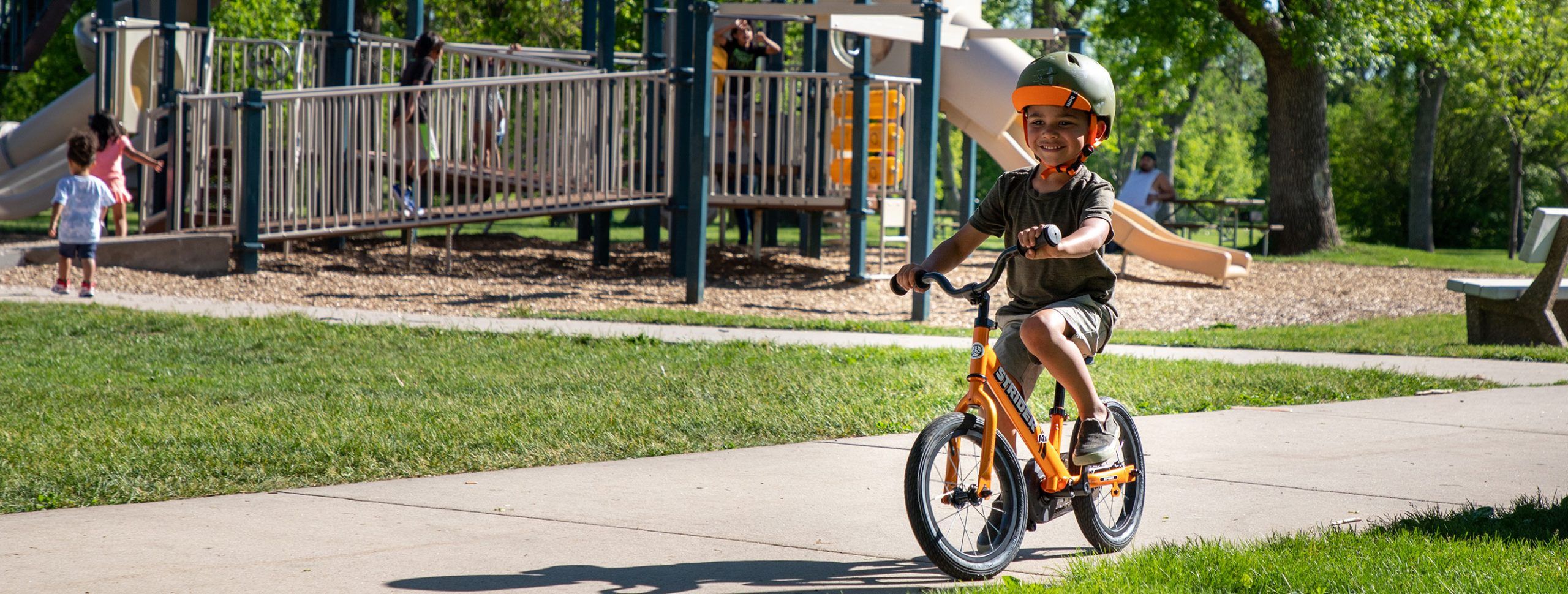 A boy confidently pedals down the sidewalk on his Tangerine Orange Strider 14x Sport bike