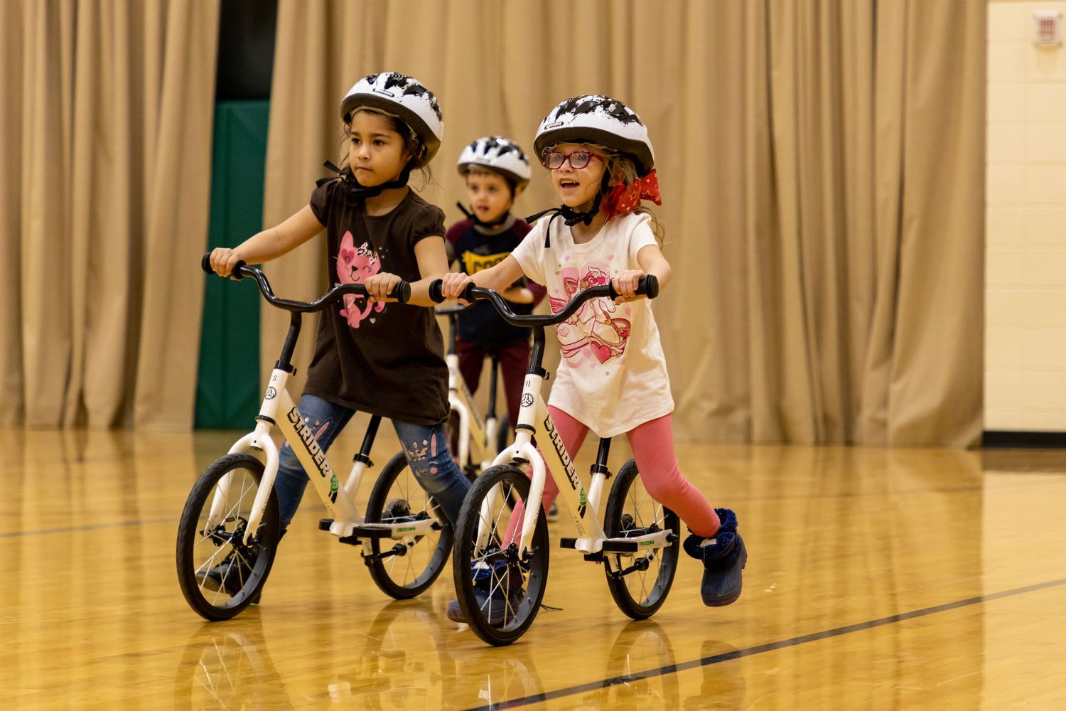 girls ride balance bikes through a school gym