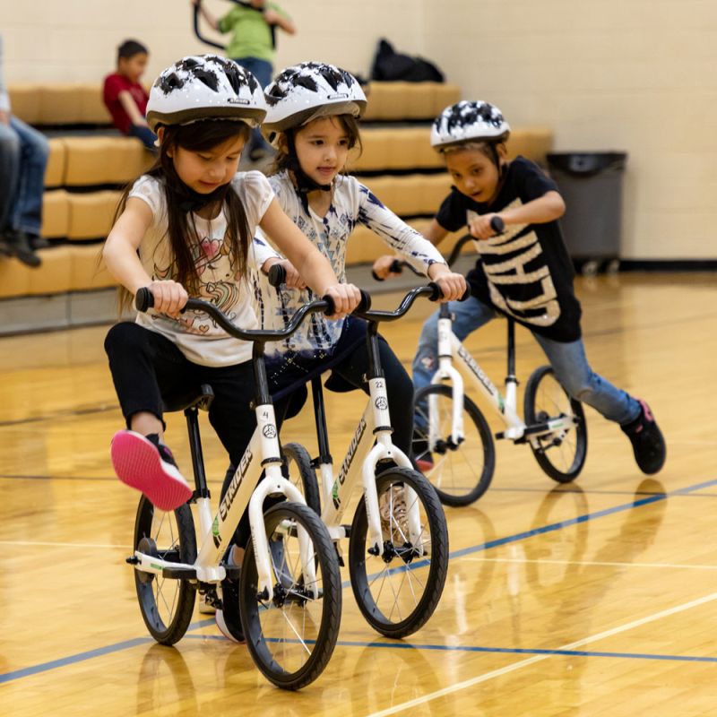 A group of children in the All Kids Bike curriculum lift their feet to "glide" on white Strider bikes
