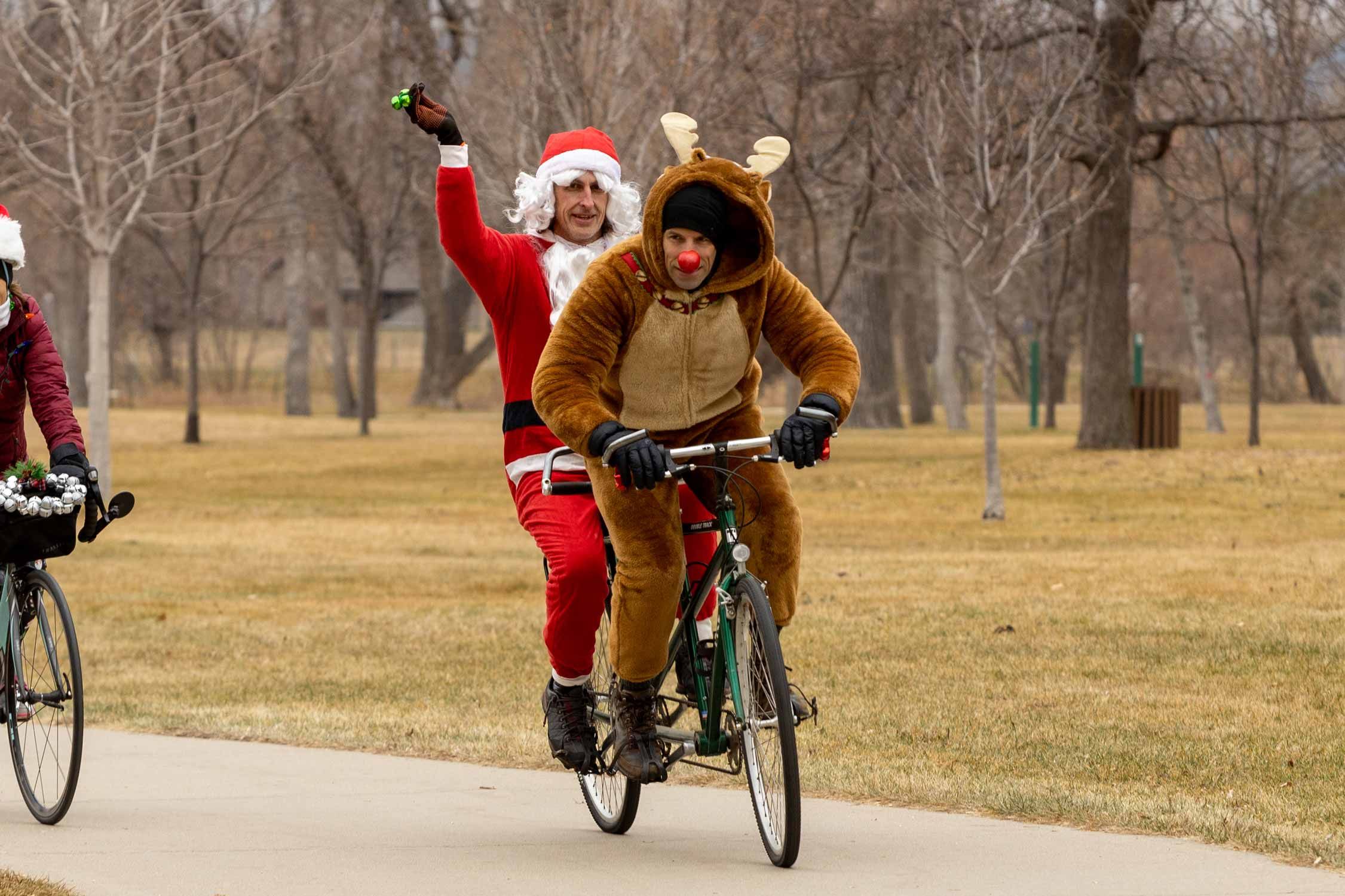 Men dressed as Santa Claus and Rudolph the Red Nosed Reindeer ride a tandem bike in a park