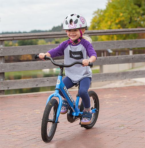A young child smiles as they pedal on their Strider 14x Bike on the sidewalk.