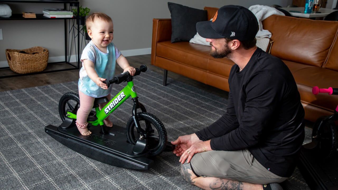 A baby on a green Rocking Bike laughs with Dad