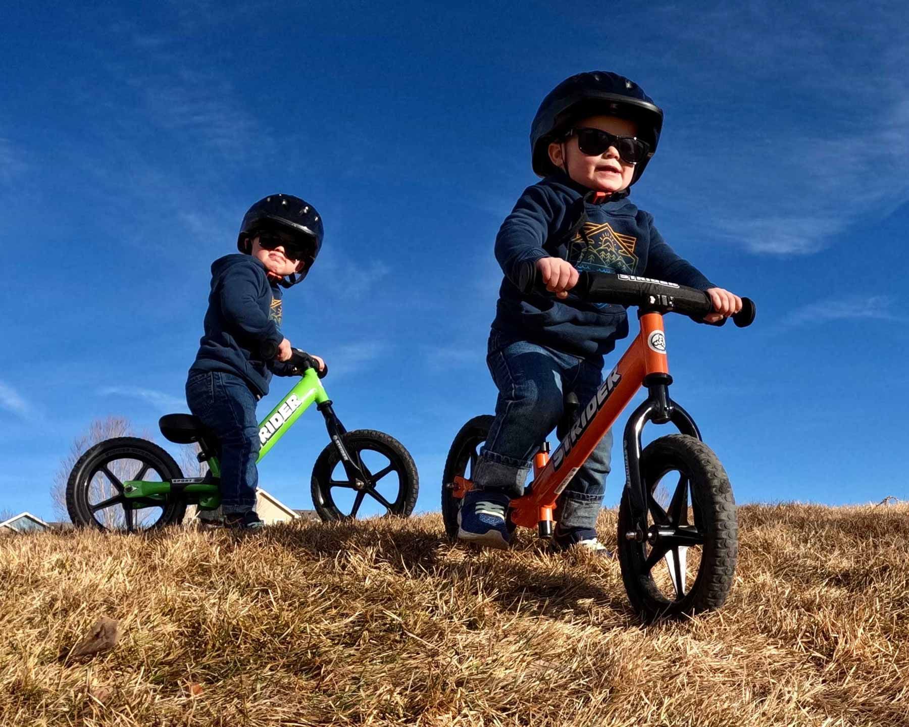 Two kids on Strider 12 Sport balance bikes prepare to go down a grassy hill.