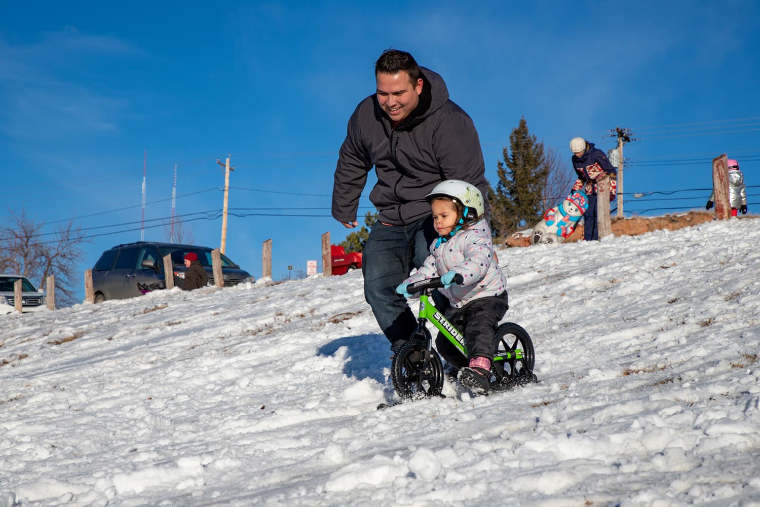 A dad runs alongside his child, who is riding a Strider bike with skis down a snowy hill
