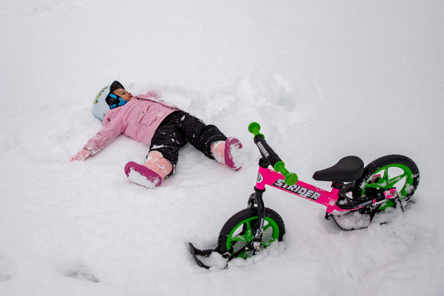 A child makes a snow angel next to a pink Strider bike with snow skis attached