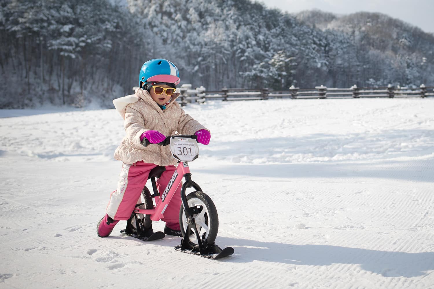 A girl rides a pink Strider bike across a flat, snowy surface