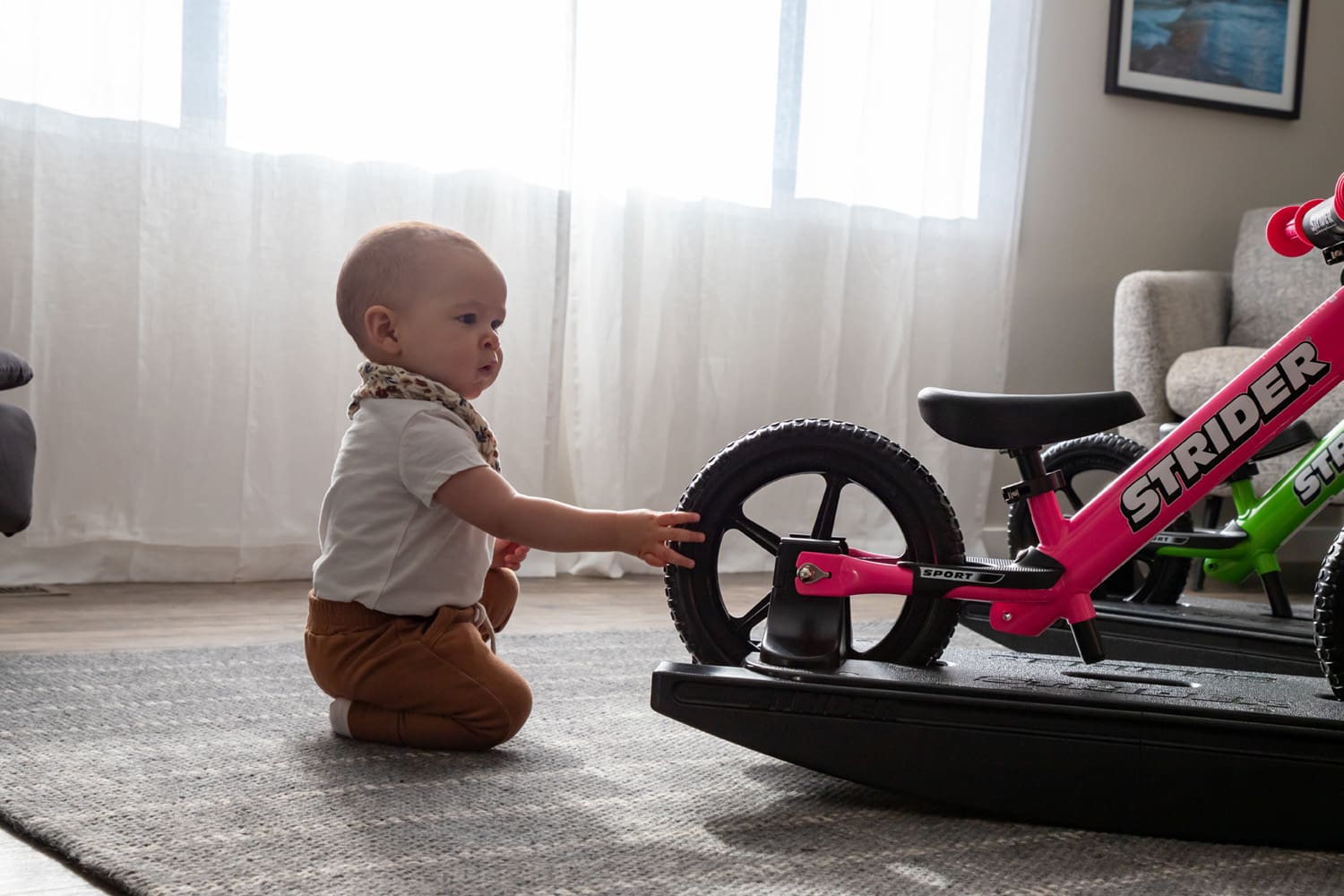 A baby checks out the wheel of a pink Strider Sport Rocking Bike