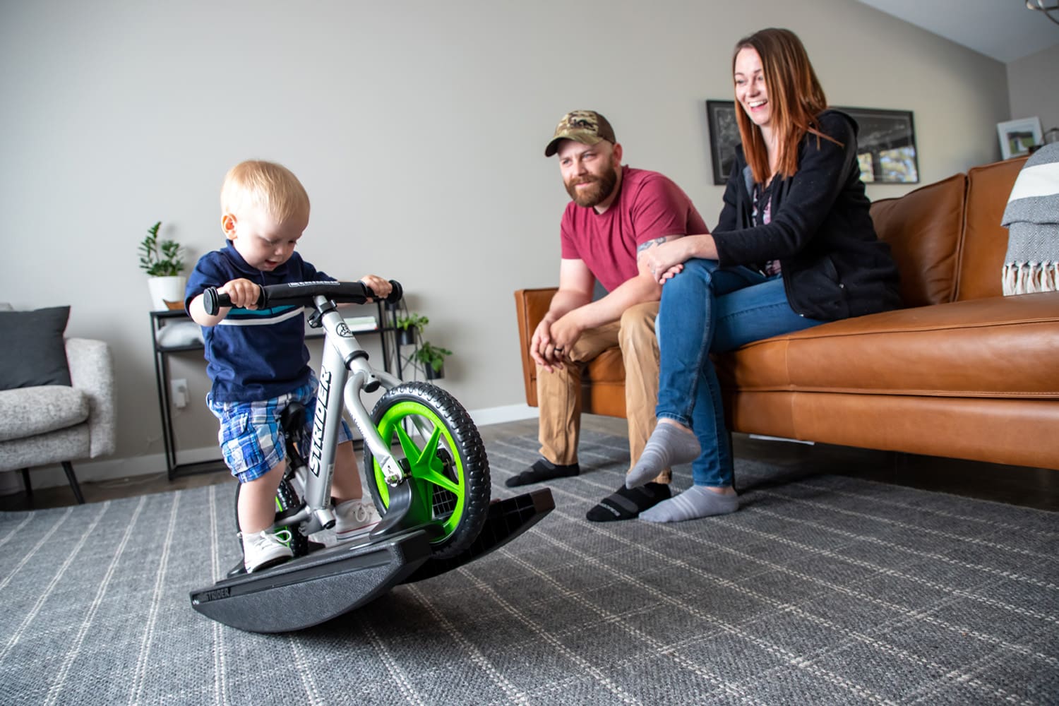 Parents watch from the sofa as their child plays on a silver Strider Pro Rocking Bike